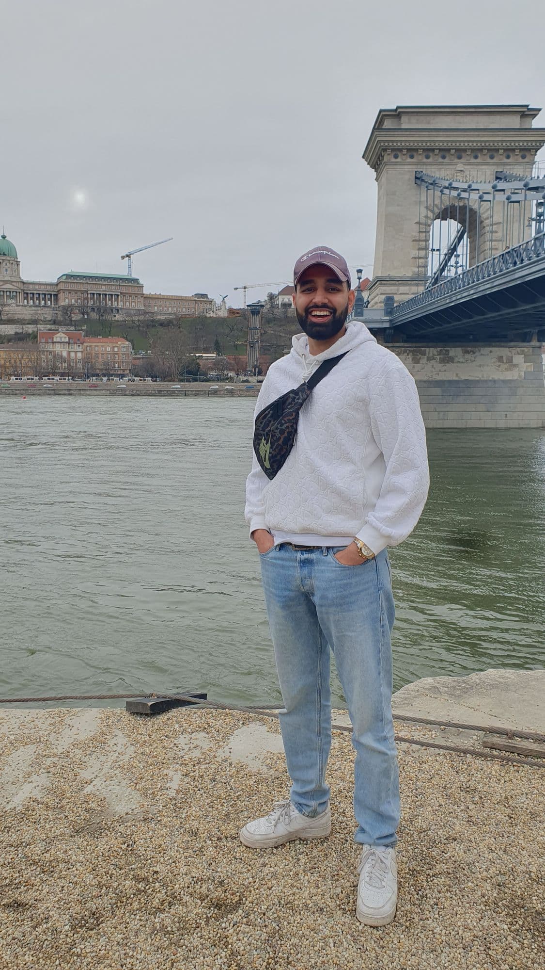 Param in a cap and white hoodie standing on the Danube embankment, the Chain Bridge beside him and Buda Castle across the river