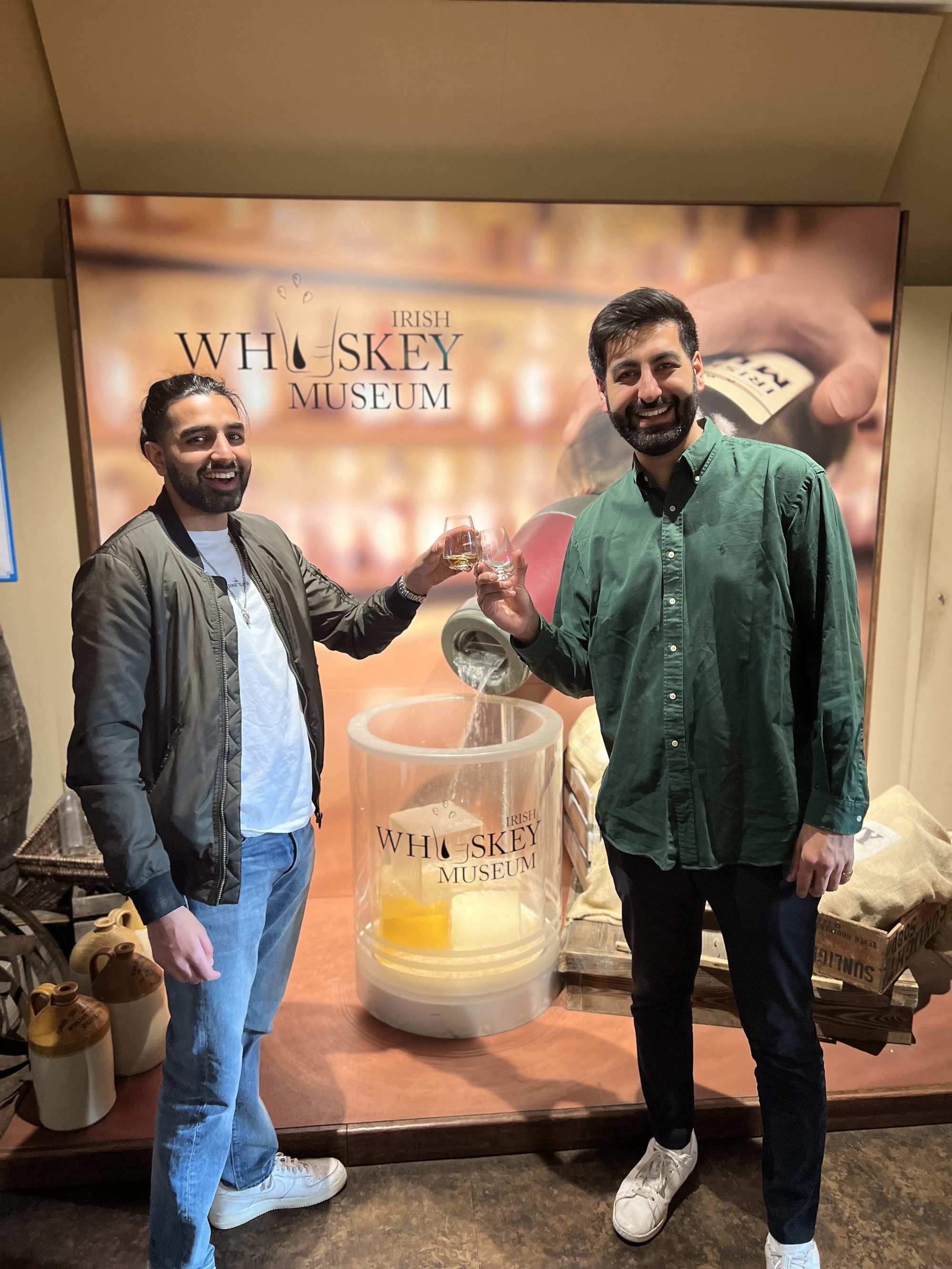 Me in an olive bomber jacket raising a small tasting glass with a man in a green shirt, in front of an Irish Whiskey Museum backdrop