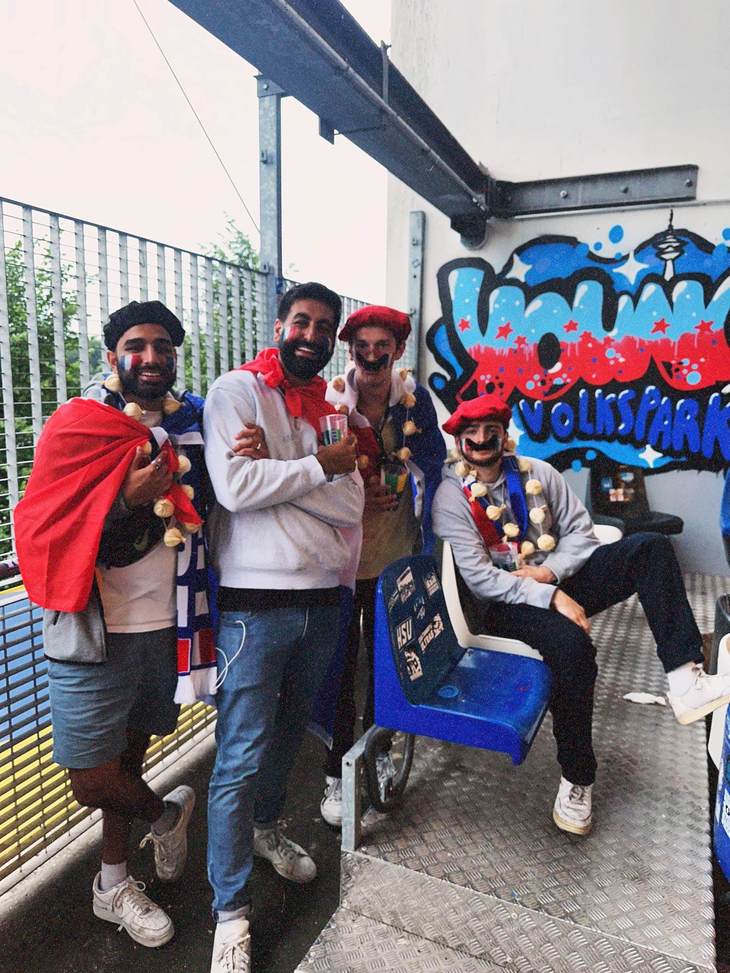 Param in a black beret and French face paint grinning under the stand at the Volksparkstadion with three friends in costume, a Volkspark graffiti wall behind