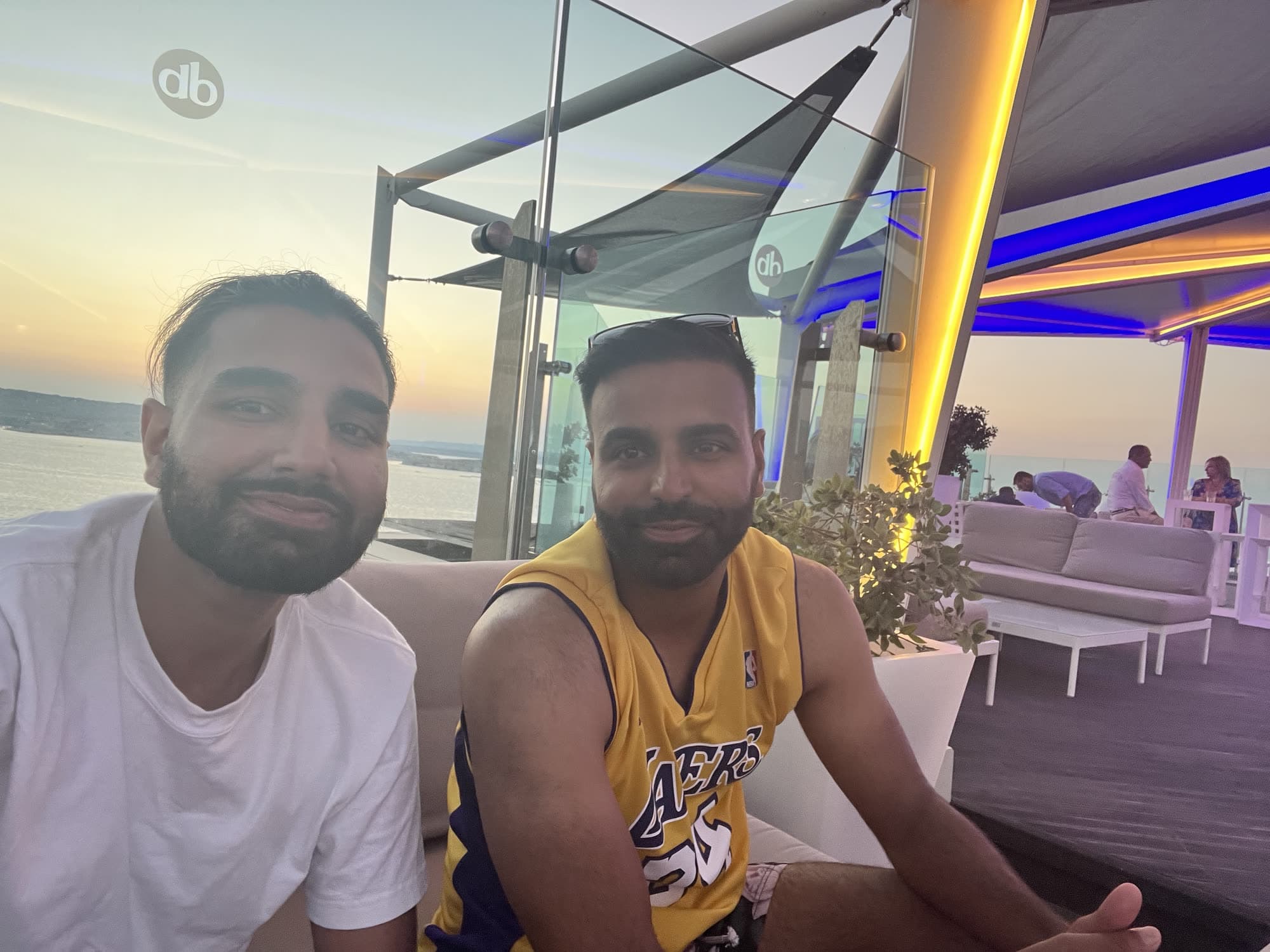 Me in a white T-shirt beside a man in a yellow Lakers jersey, both looking into the camera on a rooftop terrace at dusk, the bay behind