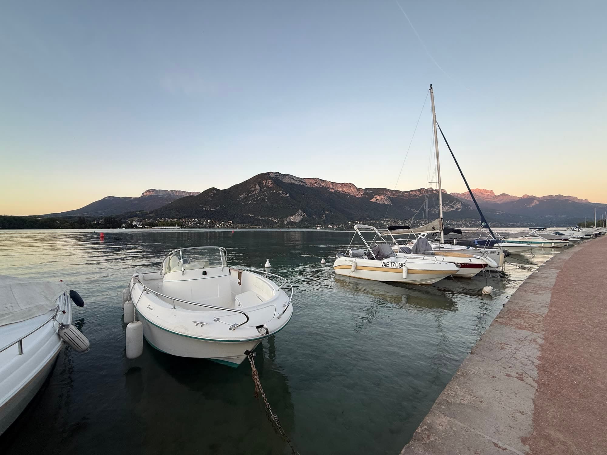 Small white motor boats and a sailing boat moored along a quay at dusk, mountains on the far shore