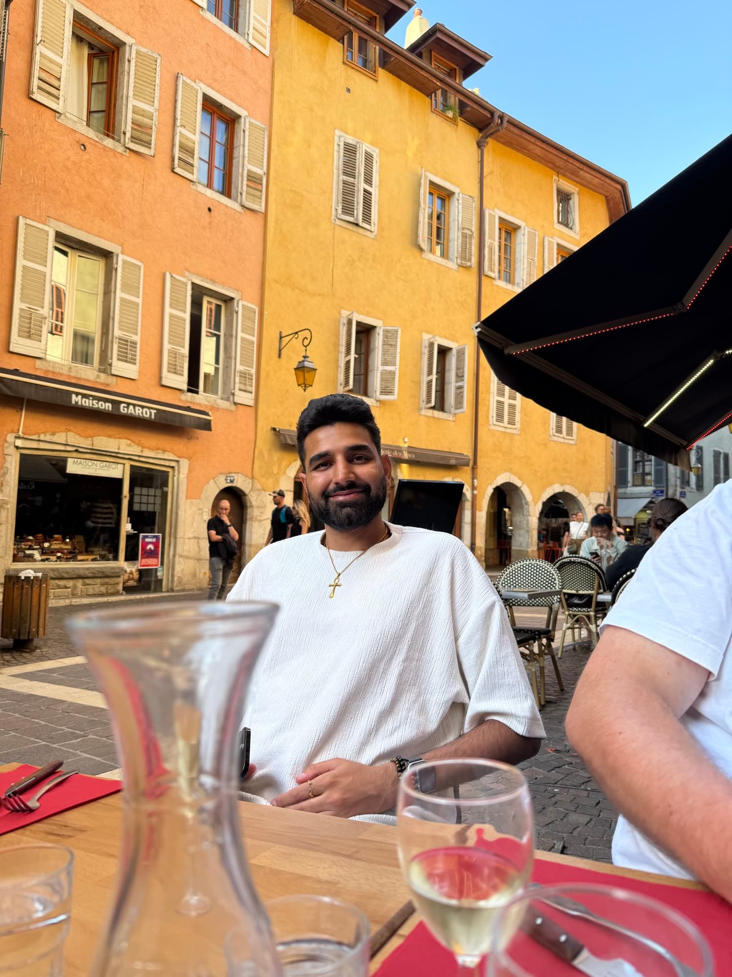 Me smiling at a café table on a square, a carafe and a glass of white wine in front of me, ochre houses behind
