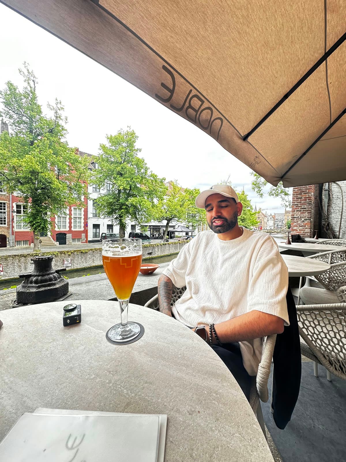 Me seated at a round stone table under an awning, a canal and brick houses behind