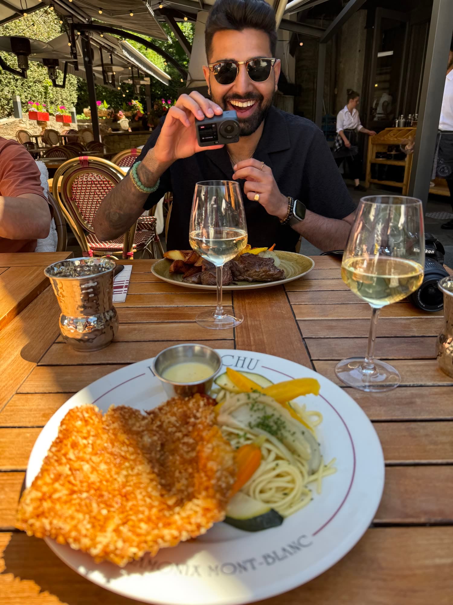 Me in sunglasses grinning on a restaurant terrace, holding up a small camera, a plate of schnitzel and two glasses of white wine in front of me