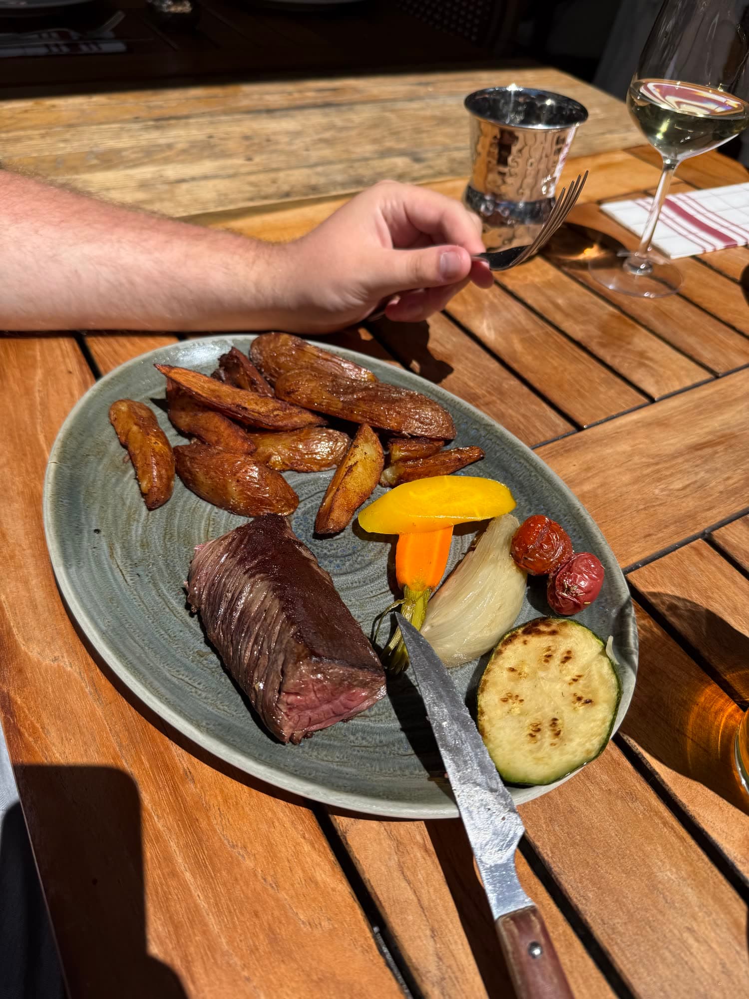 A plate of steak, roast potatoes and grilled vegetables on a wooden table in the sun