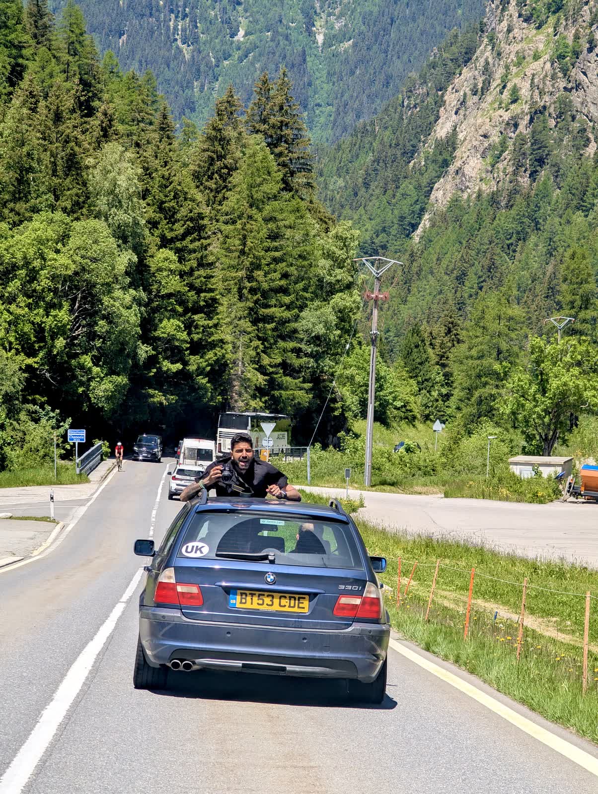 Me half out of the sunroof of the blue BMW estate ahead with a camera in my hands, pine forest and a lorry behind