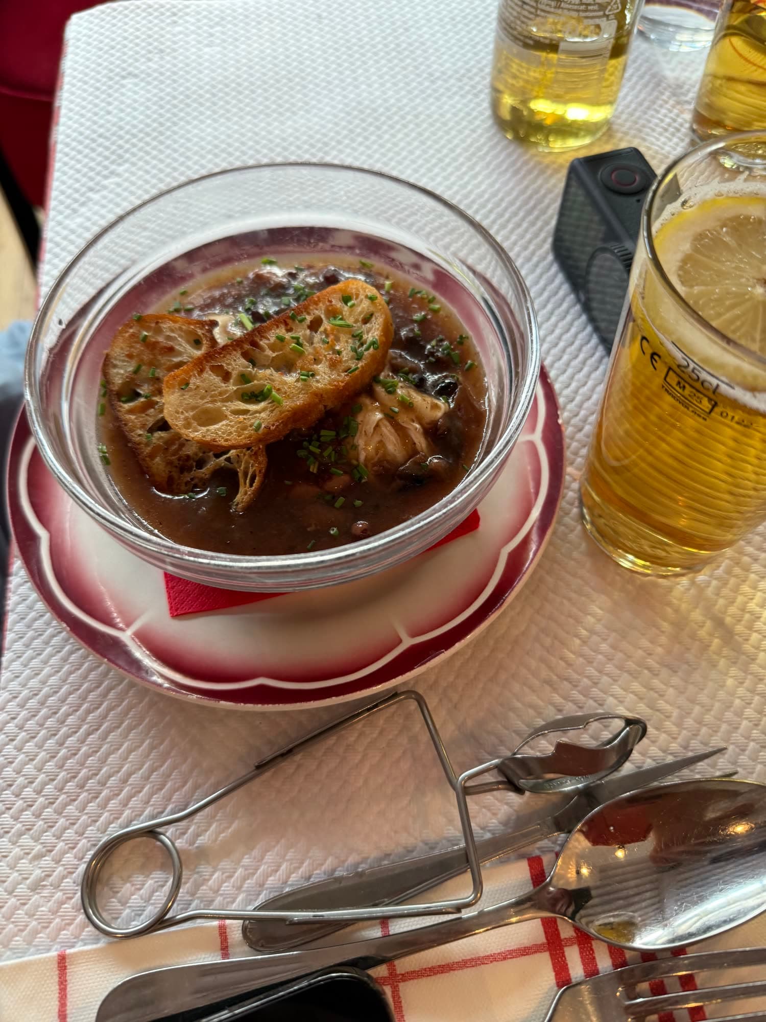 French onion soup under a crust of bread and melted cheese in a glass bowl, a beer beside it