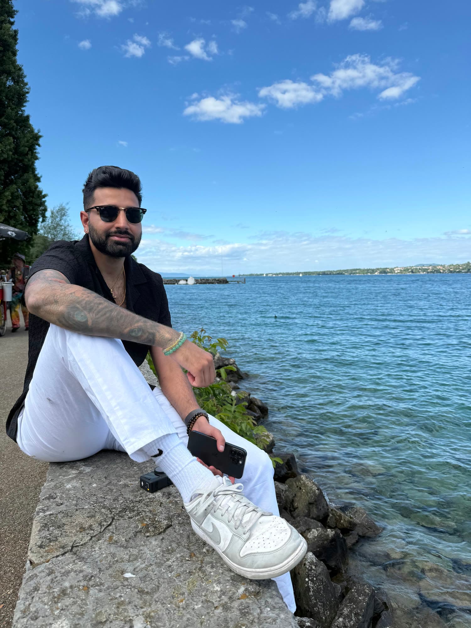 Me in sunglasses, a black shirt and white trousers, sitting on a stone wall at the edge of the lake