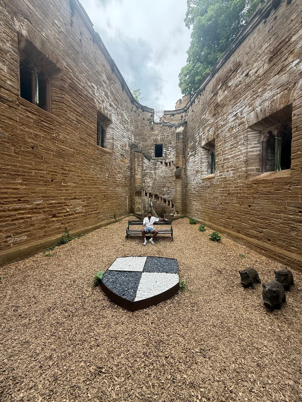 A narrow sandstone yard with a black and white quartered shield in the gravel and me on a bench at the far end
