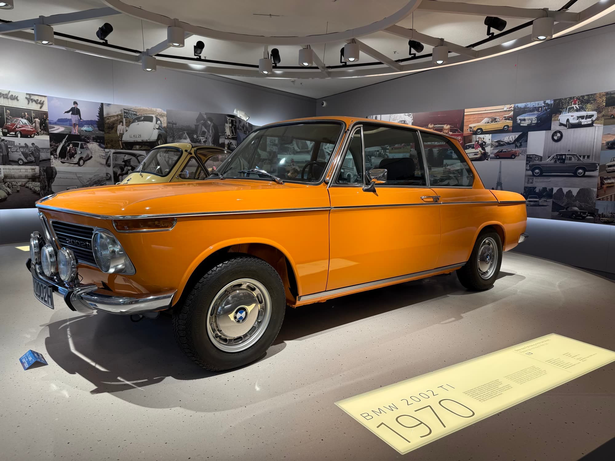 An orange 1970 BMW 2002 ti on a round plinth, the year marked on the floor in front of it