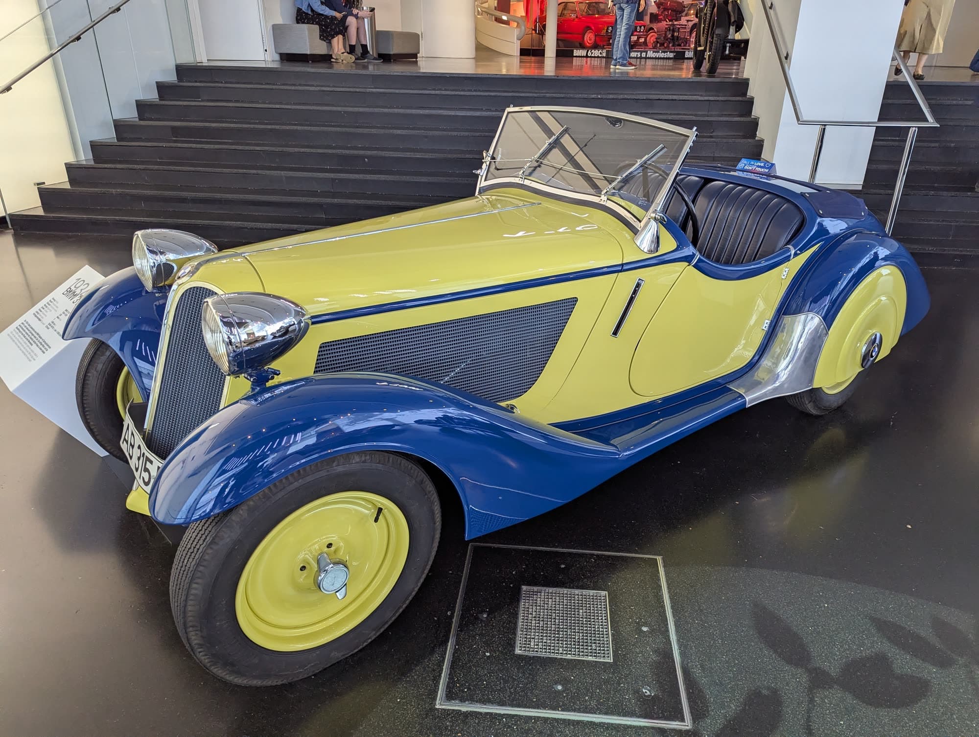 A 1930s BMW roadster in yellow with blue wings and yellow wheels on the museum floor