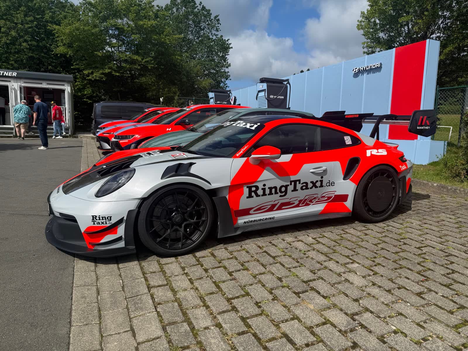 A Porsche 911 GT3 RS in red and white Ring Taxi livery parked on cobbles in the paddock