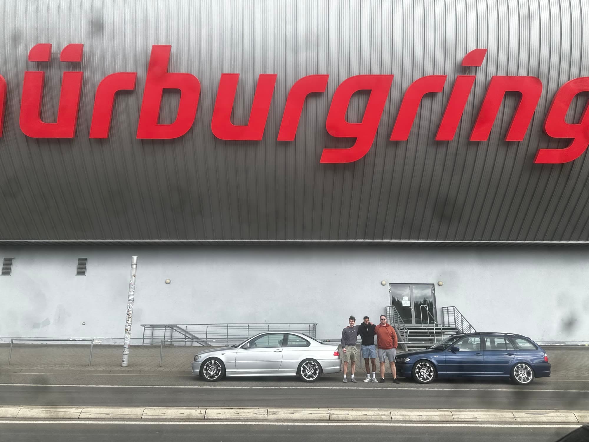 The silver coupe and the blue estate parked beneath the red nürburgring lettering on the grandstand wall, three of us standing between them