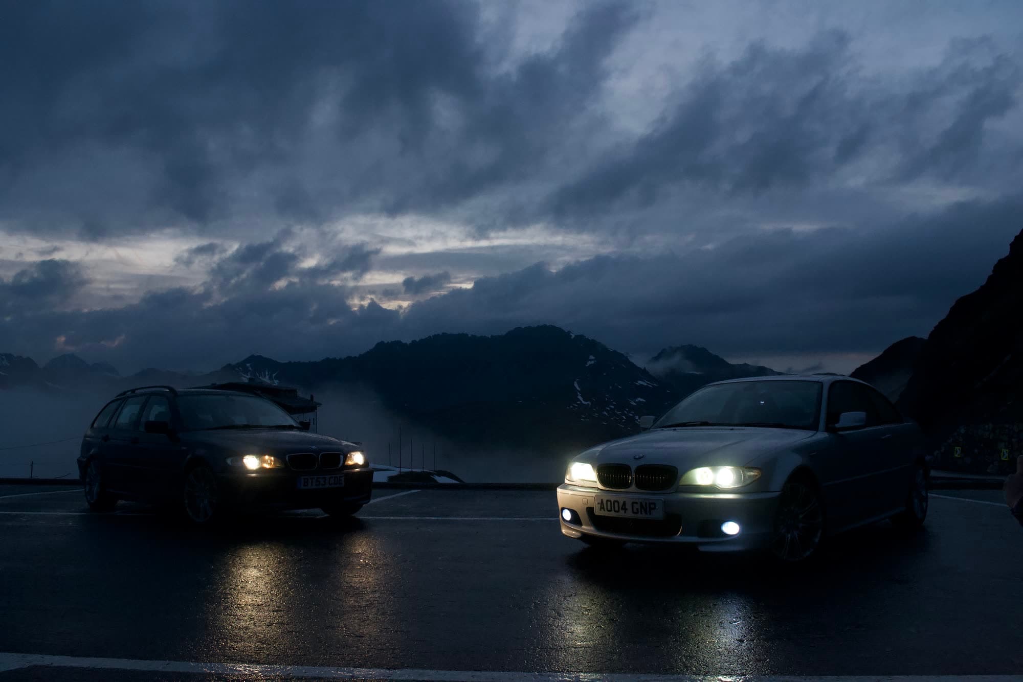 The two BMWs at the Stelvio summit at dusk, headlights on, cloud over the peaks behind
