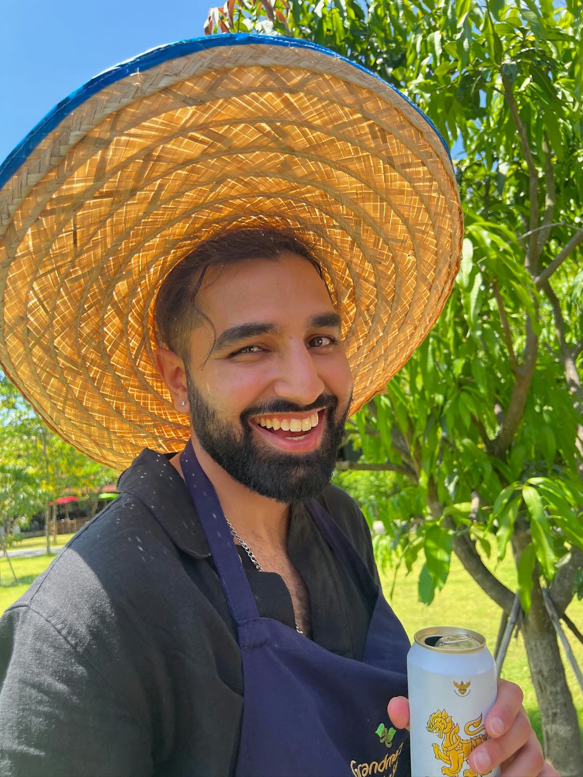 Param laughing under a wide woven straw hat, in a black shirt and a dark apron, a cold can in his hand and a tree behind him