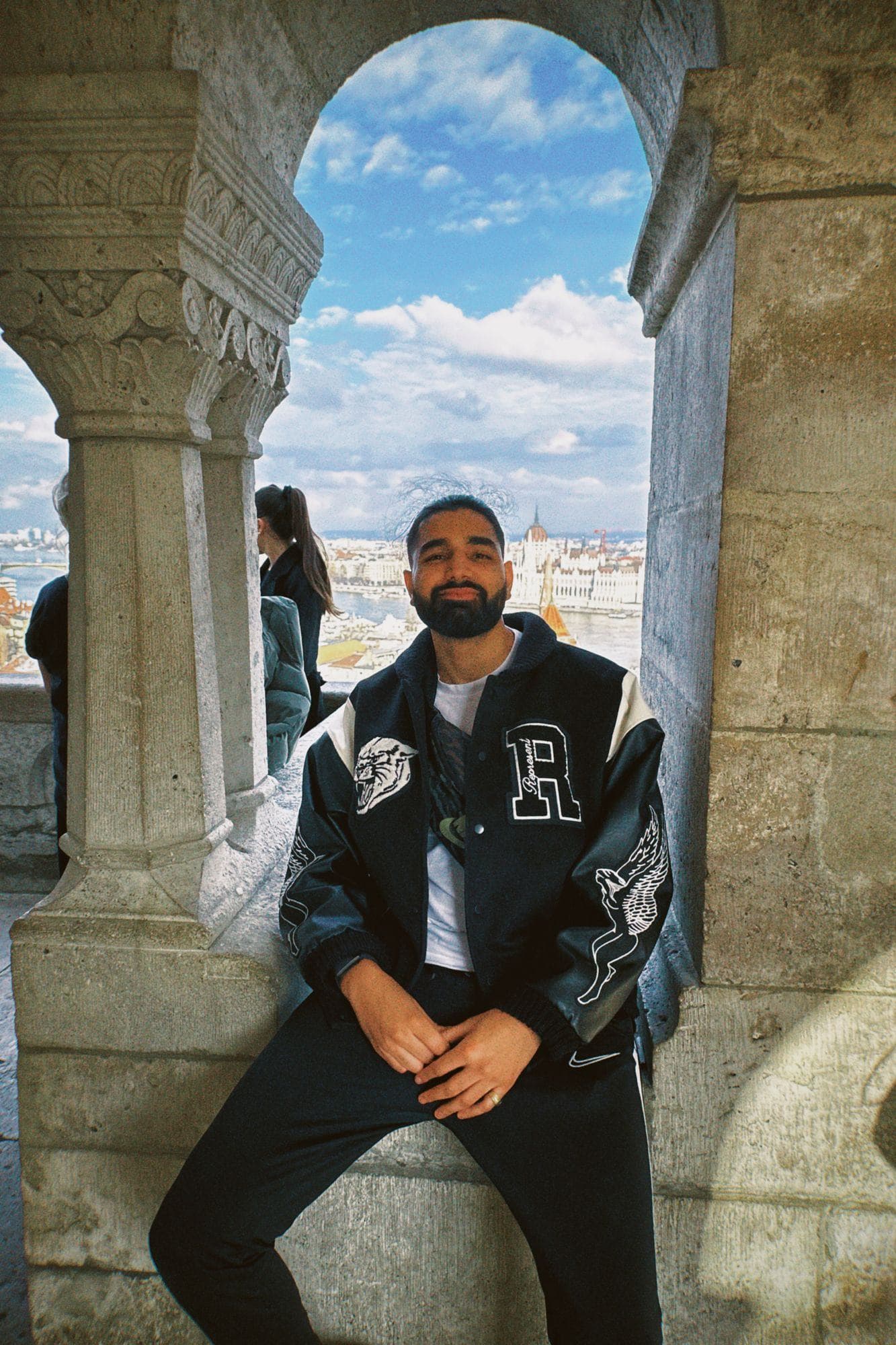 Param sitting in a stone arch at Fisherman’s Bastion in a varsity jacket, blue sky and the city beyond
