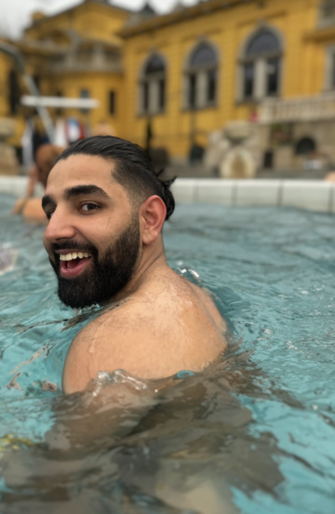 Param laughing over his shoulder in the warm pool at Széchenyi, the yellow facade blurred behind him