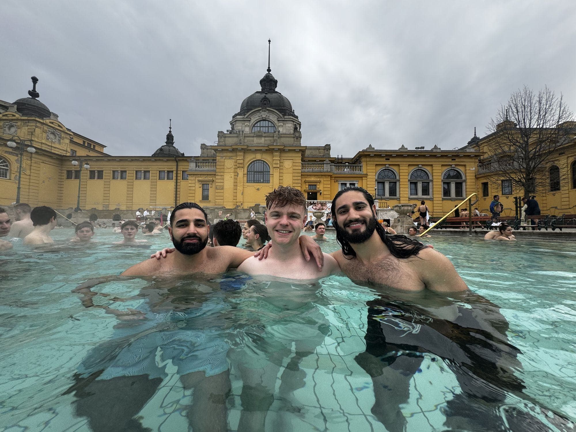 Param and two friends waist-deep in the outdoor pool at Széchenyi Baths, the yellow bathhouse and its dome behind them