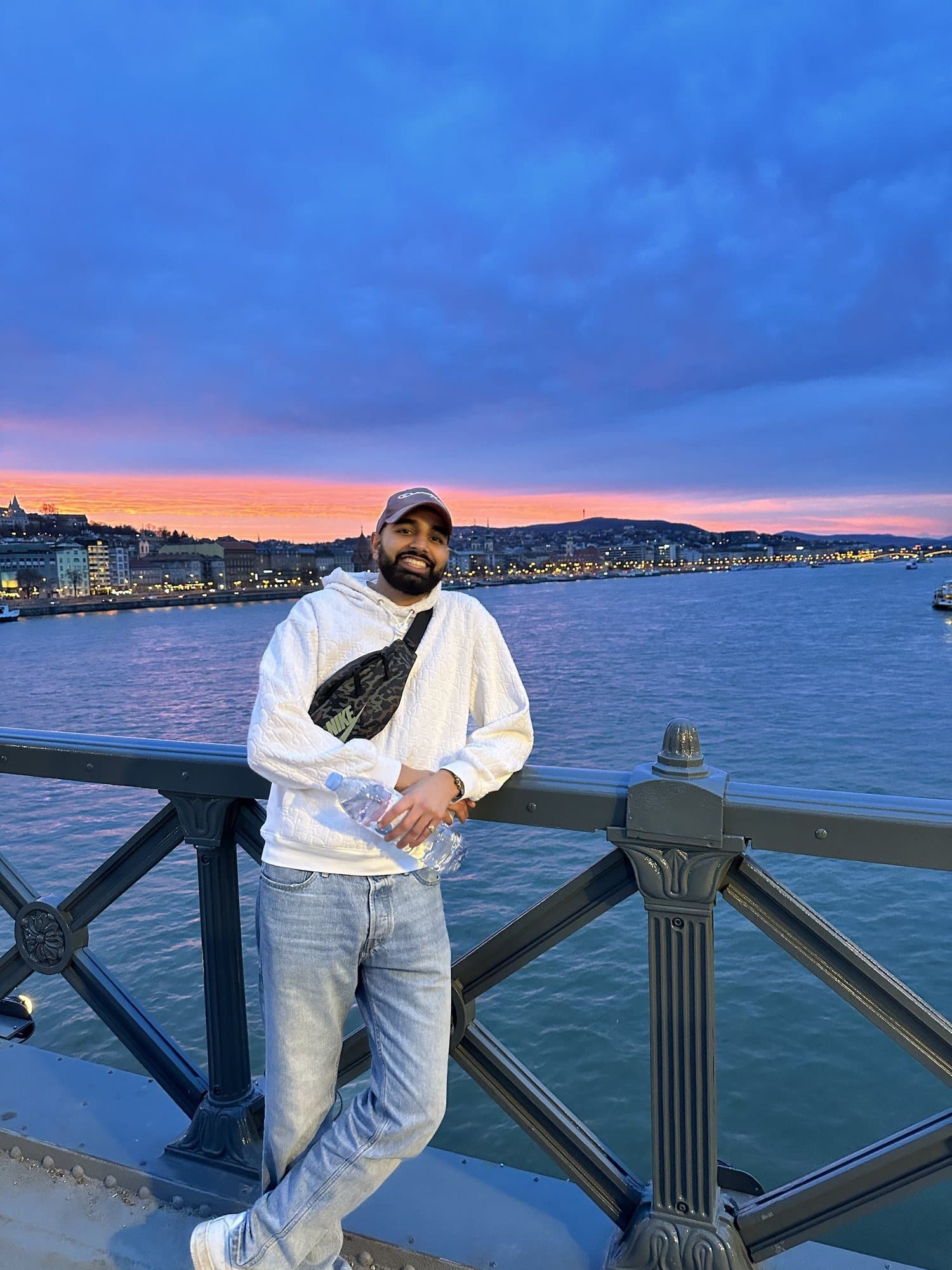 Param leaning on the iron railing of the Chain Bridge at dusk, a pink sunset over the lit Buda bank behind him
