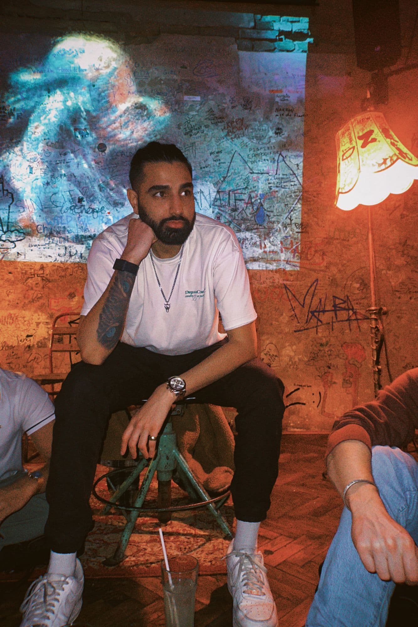 Param sitting on a green metal stool in a ruin bar, chin on his fist, a lit lampshade and graffiti-covered walls behind him