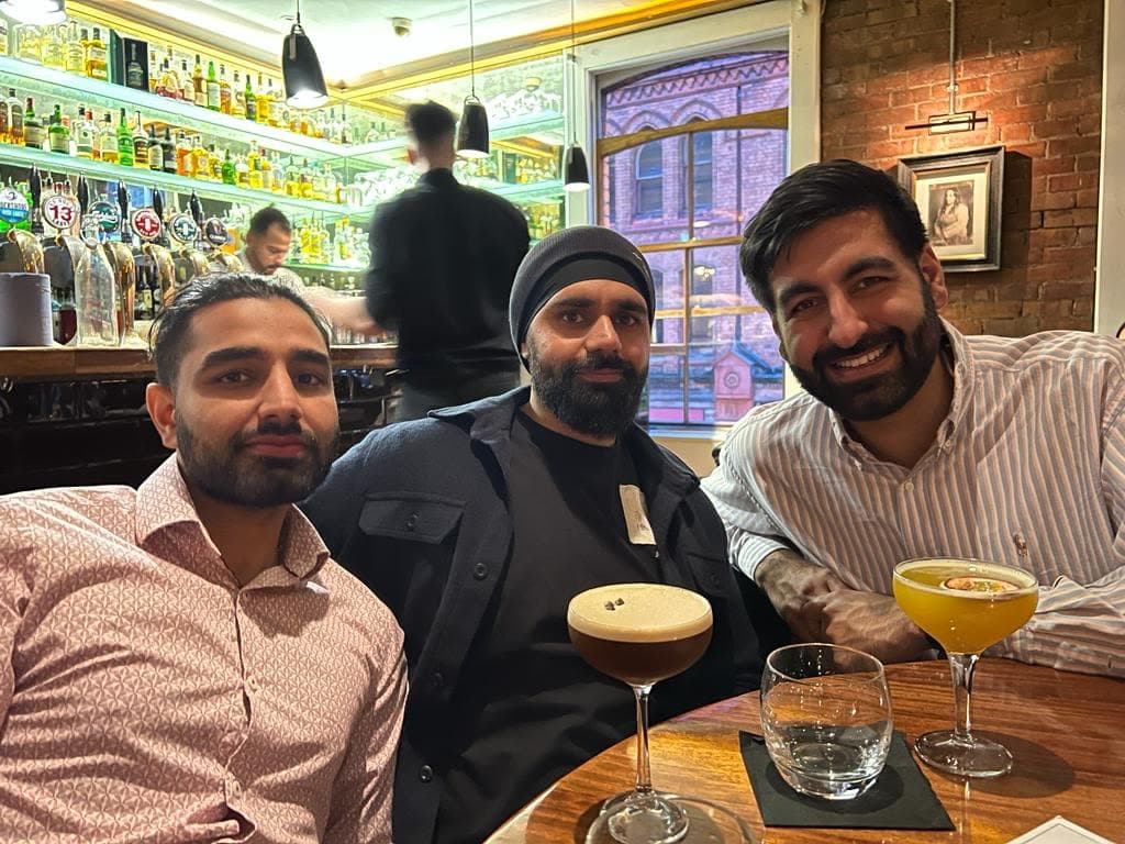 Me on the left in a pink patterned shirt at a bar table with two others, an espresso martini and a cocktail on the table, the back bar lit behind