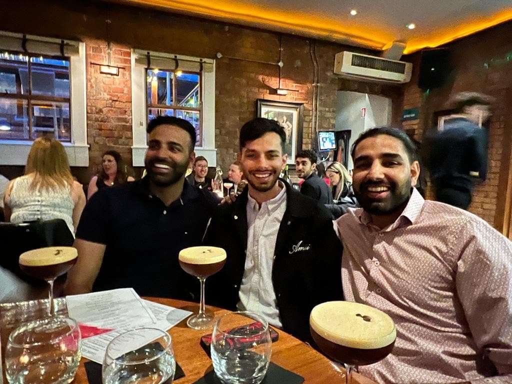 Me on the right in a pink patterned shirt, smiling beside two others at a round table with espresso martinis, a brick wall behind