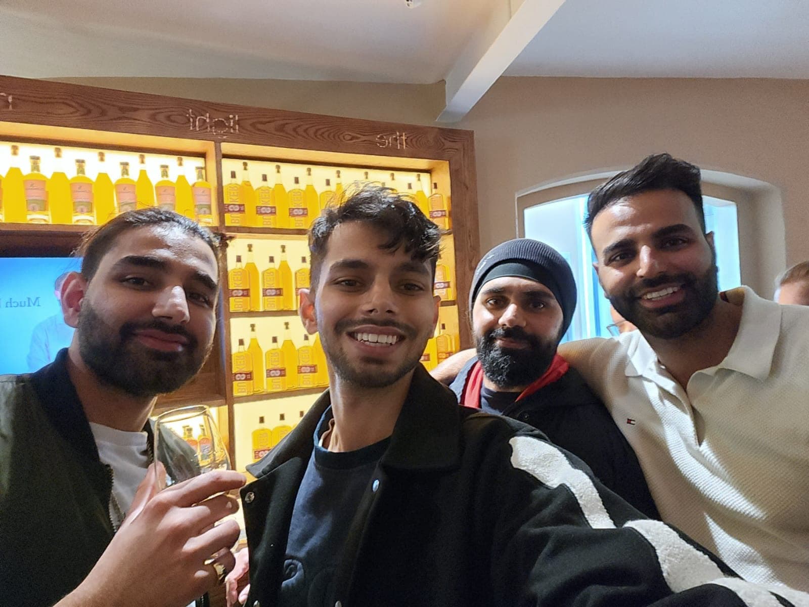 Me on the left holding a tasting glass in a four-person selfie, shelves of whiskey bottles lit yellow behind