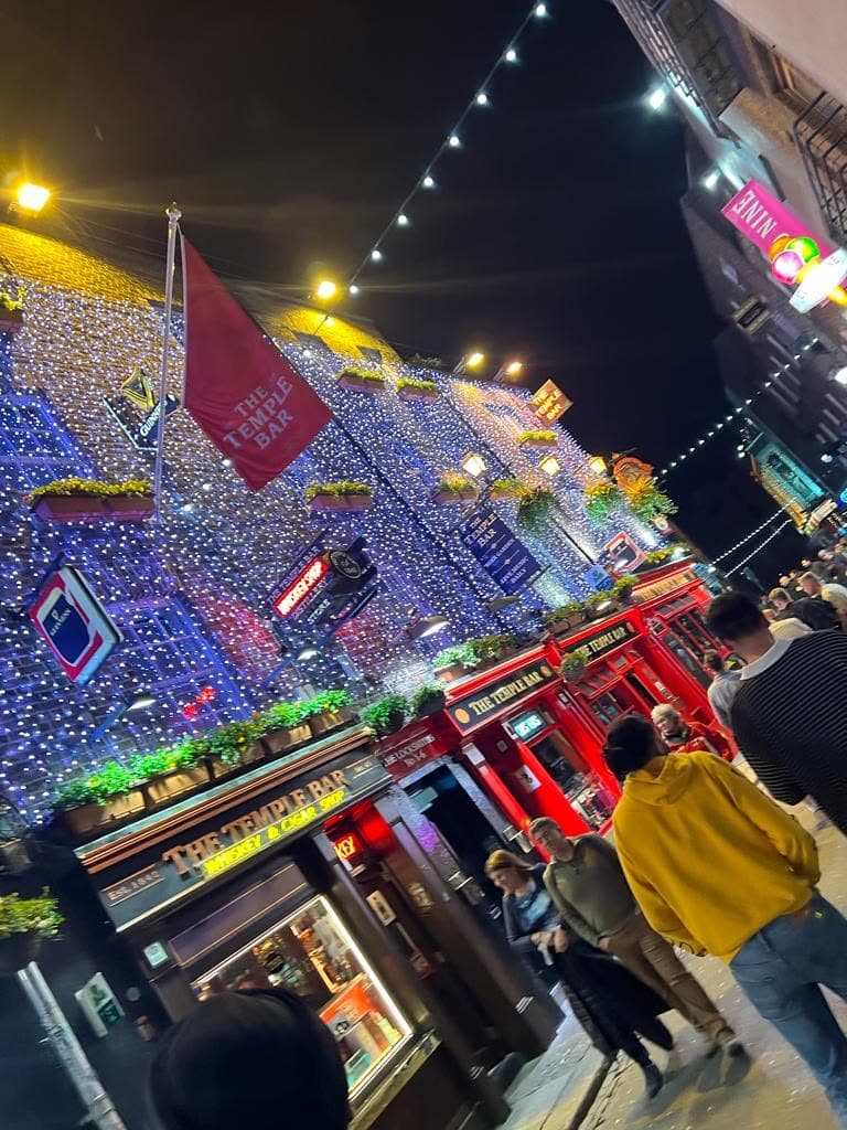 A night street in Temple Bar with a pub front covered in fairy lights, me from behind in a yellow hoodie walking with the crowd