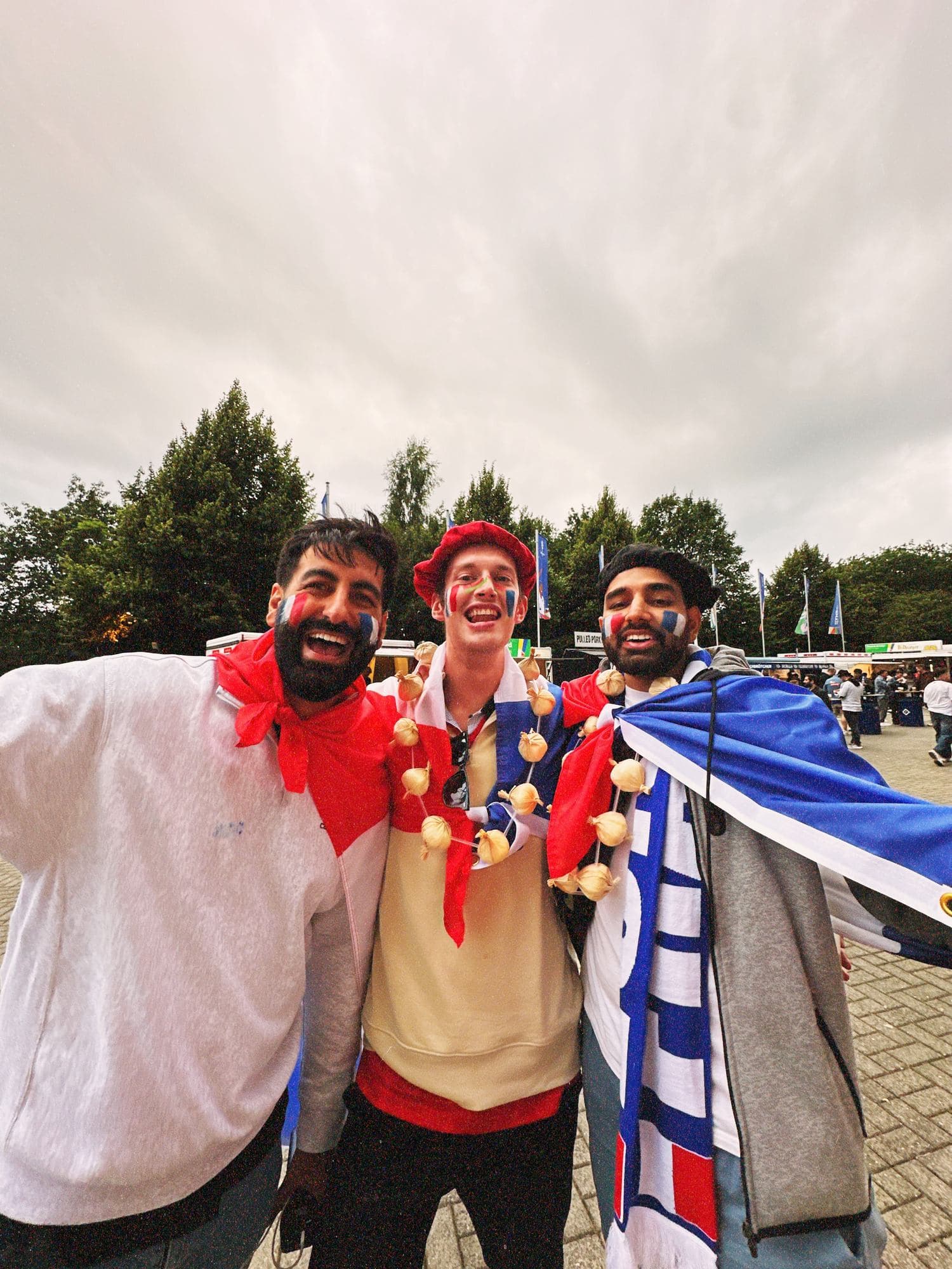 Param cheering in a beret and face paint, wrapped in a French flag, between two friends in the grounds outside the stadium