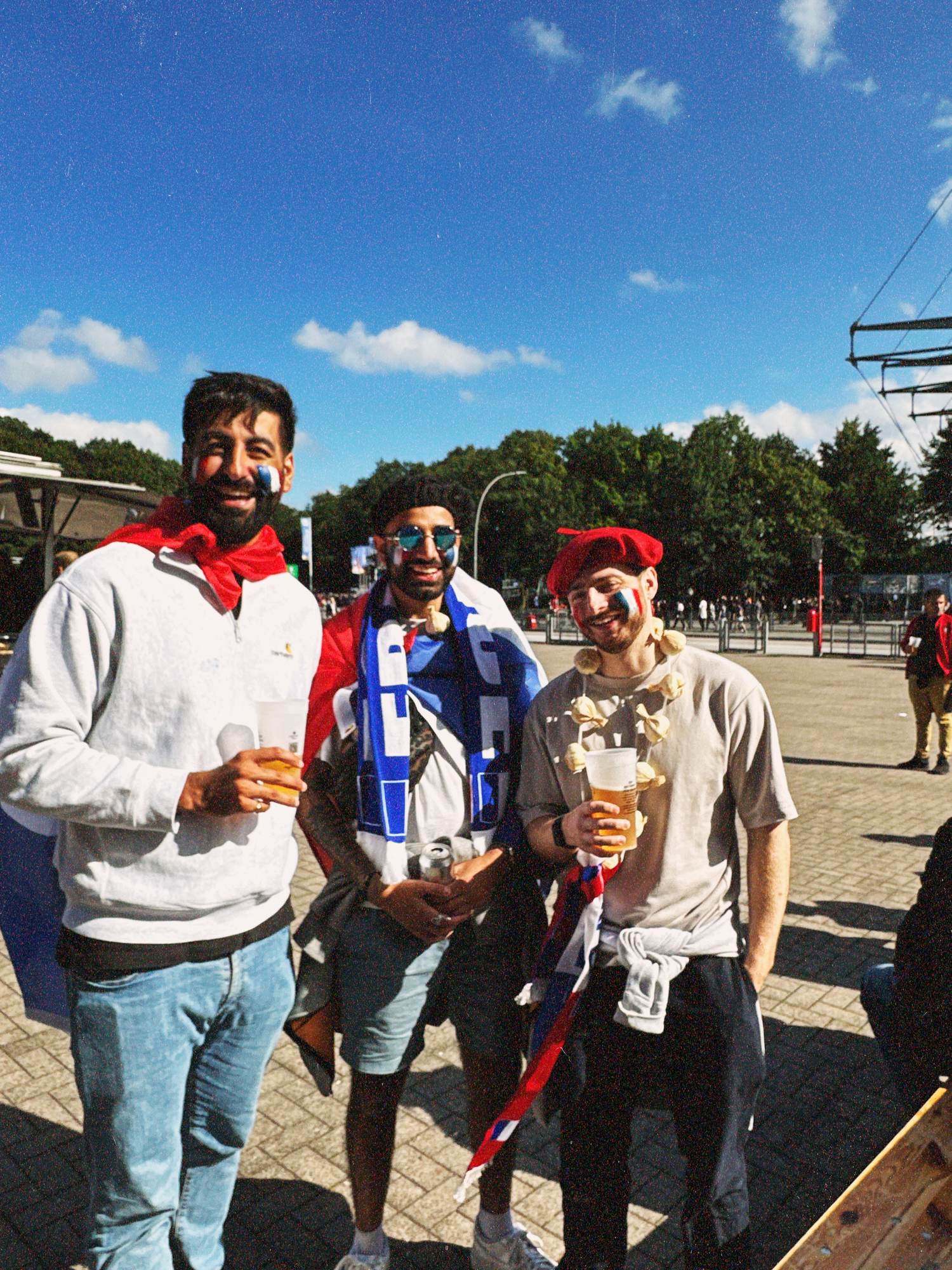 Param in round mirrored sunglasses and a France scarf smiling between two friends holding beers on a sunlit forecourt