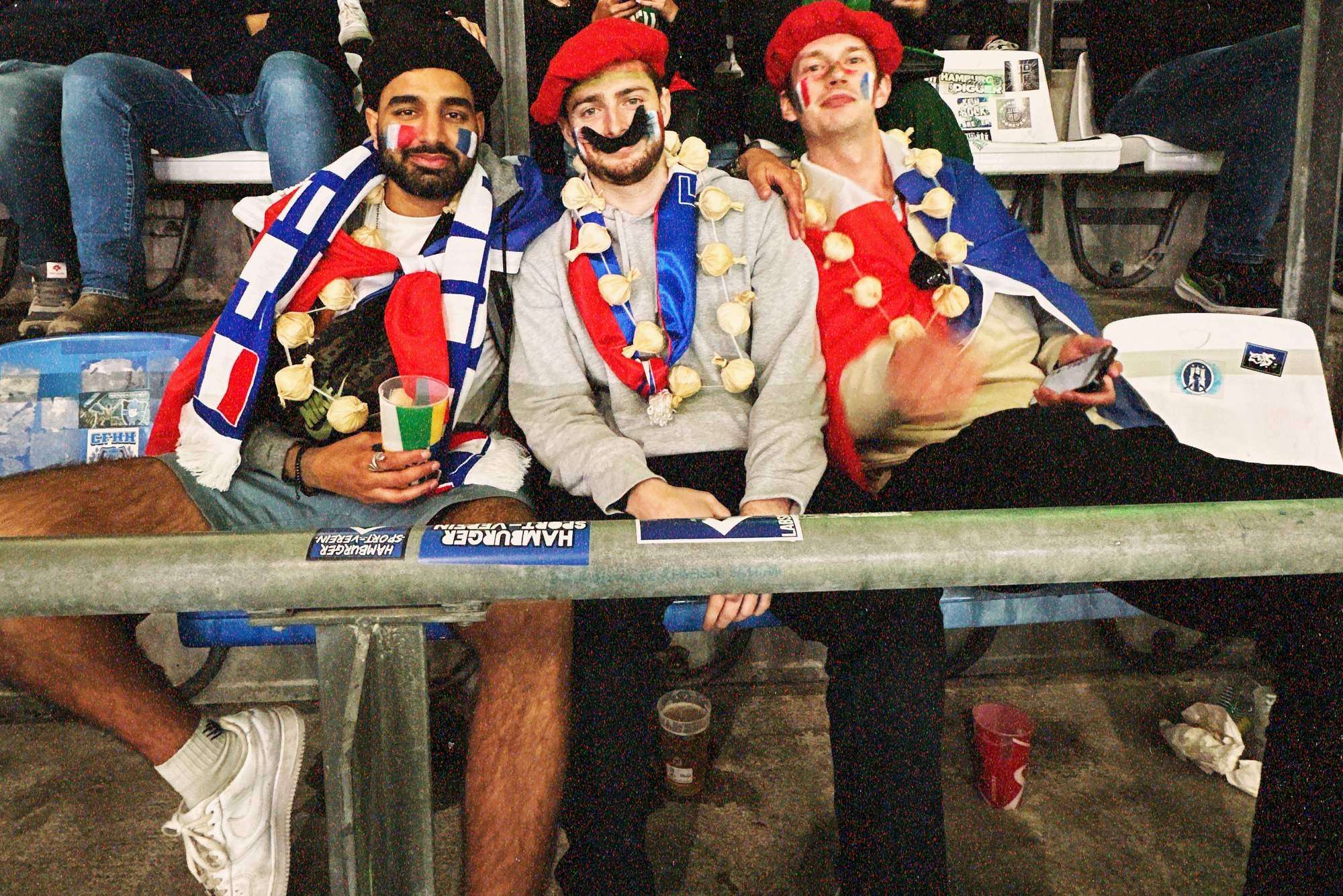 Param seated in the stand with two friends in red berets and painted moustaches, all three in garlic necklaces