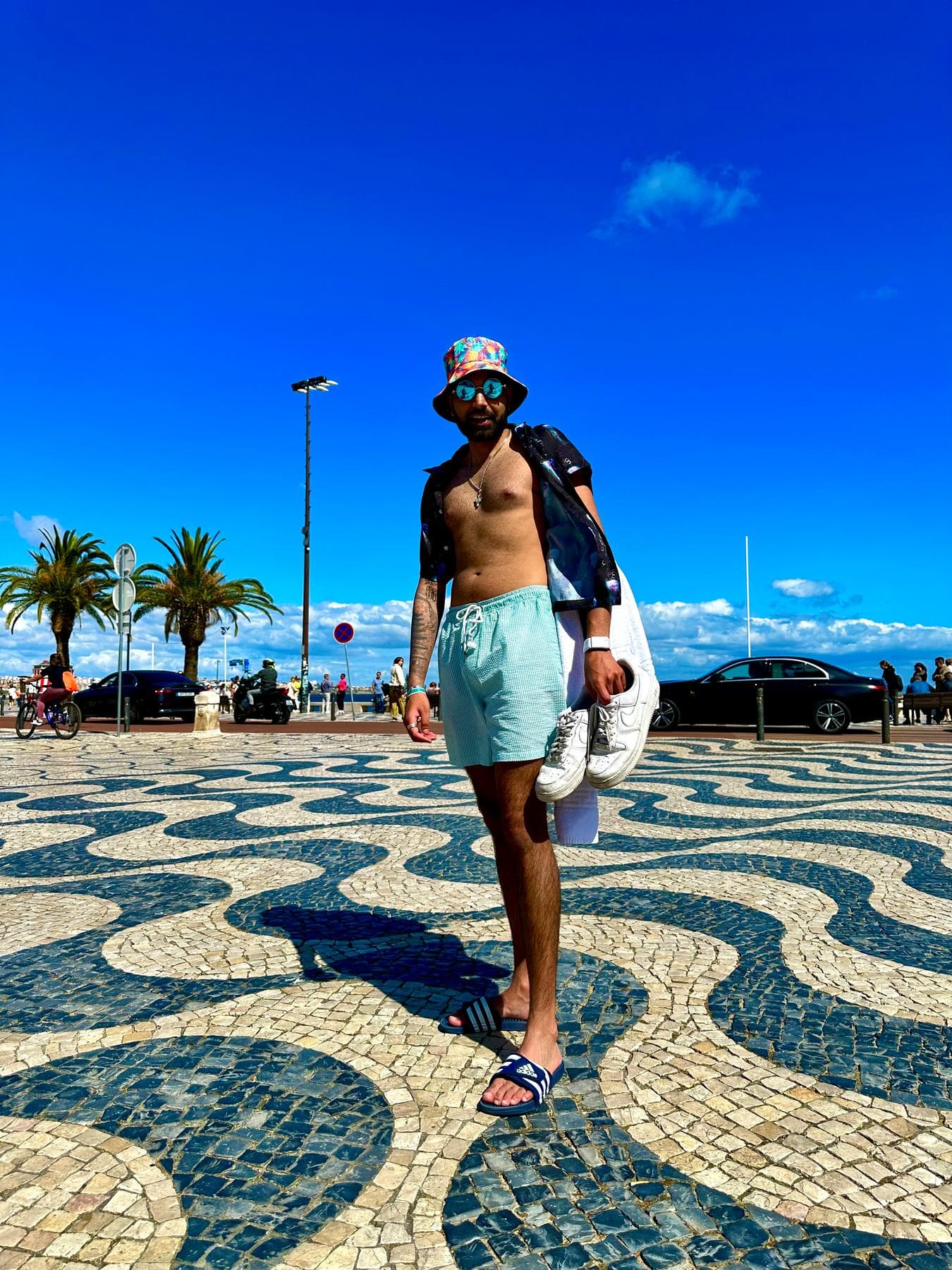 Param in a bucket hat and swim shorts walking across black-and-white wave-patterned cobbles by the sea
