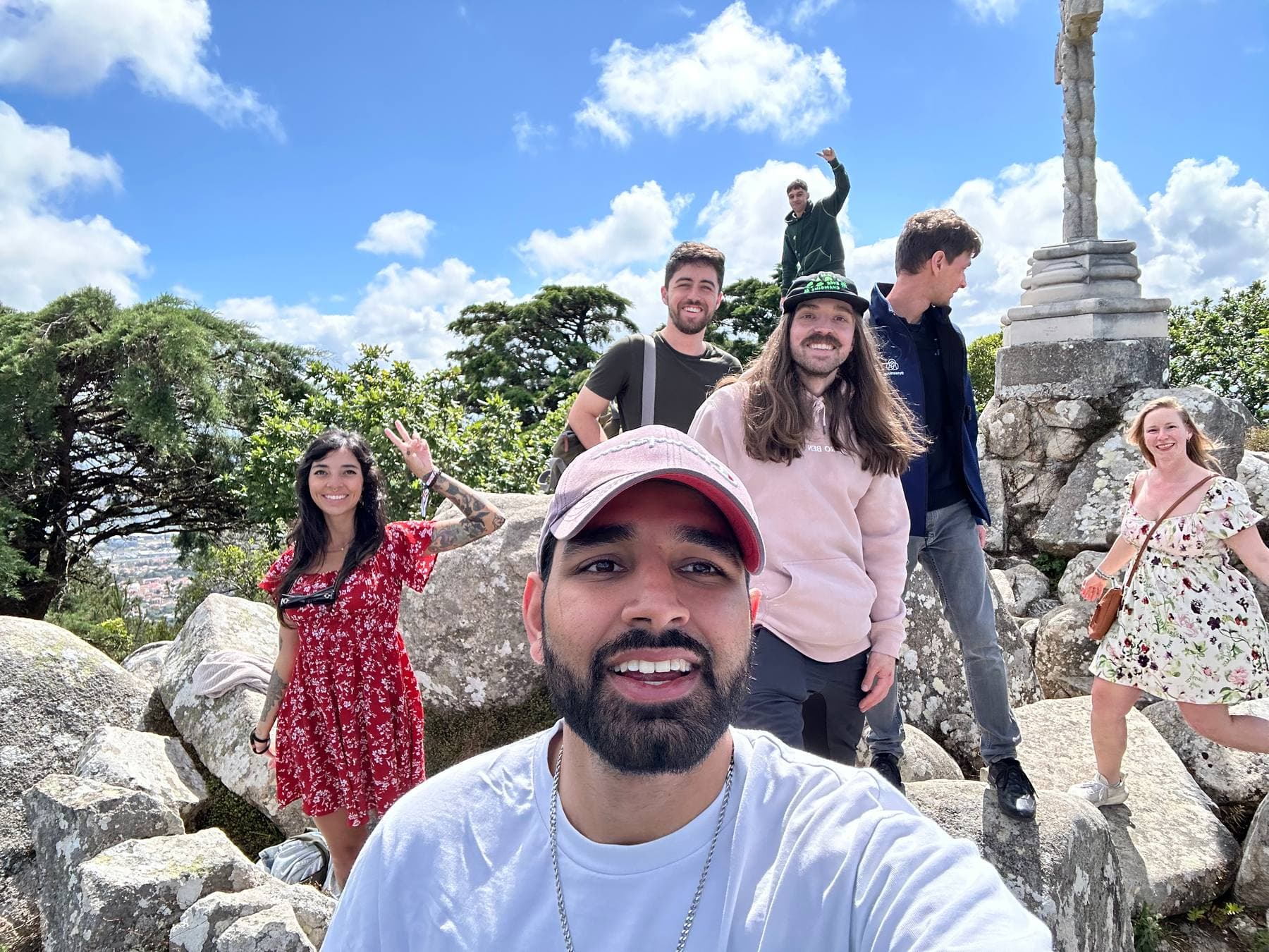 Param grinning in a selfie on a rocky summit with a group of friends and a stone cross behind them