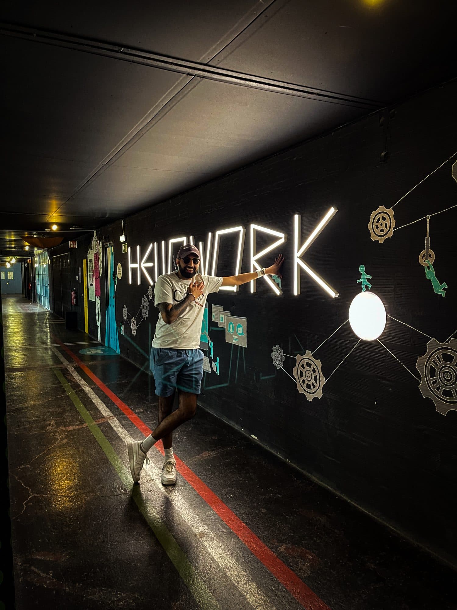 Param smiling in sunglasses and a cap, one hand on his chest and one on a lit neon sign along a dark corridor wall