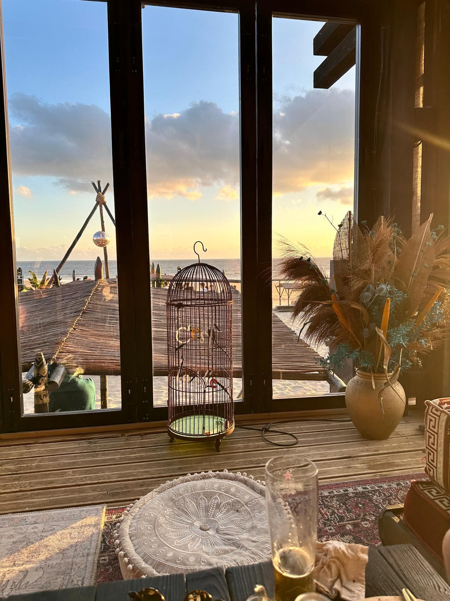 Evening light over the sea through the open frame of a beach bar, a birdcage and a glass on the table