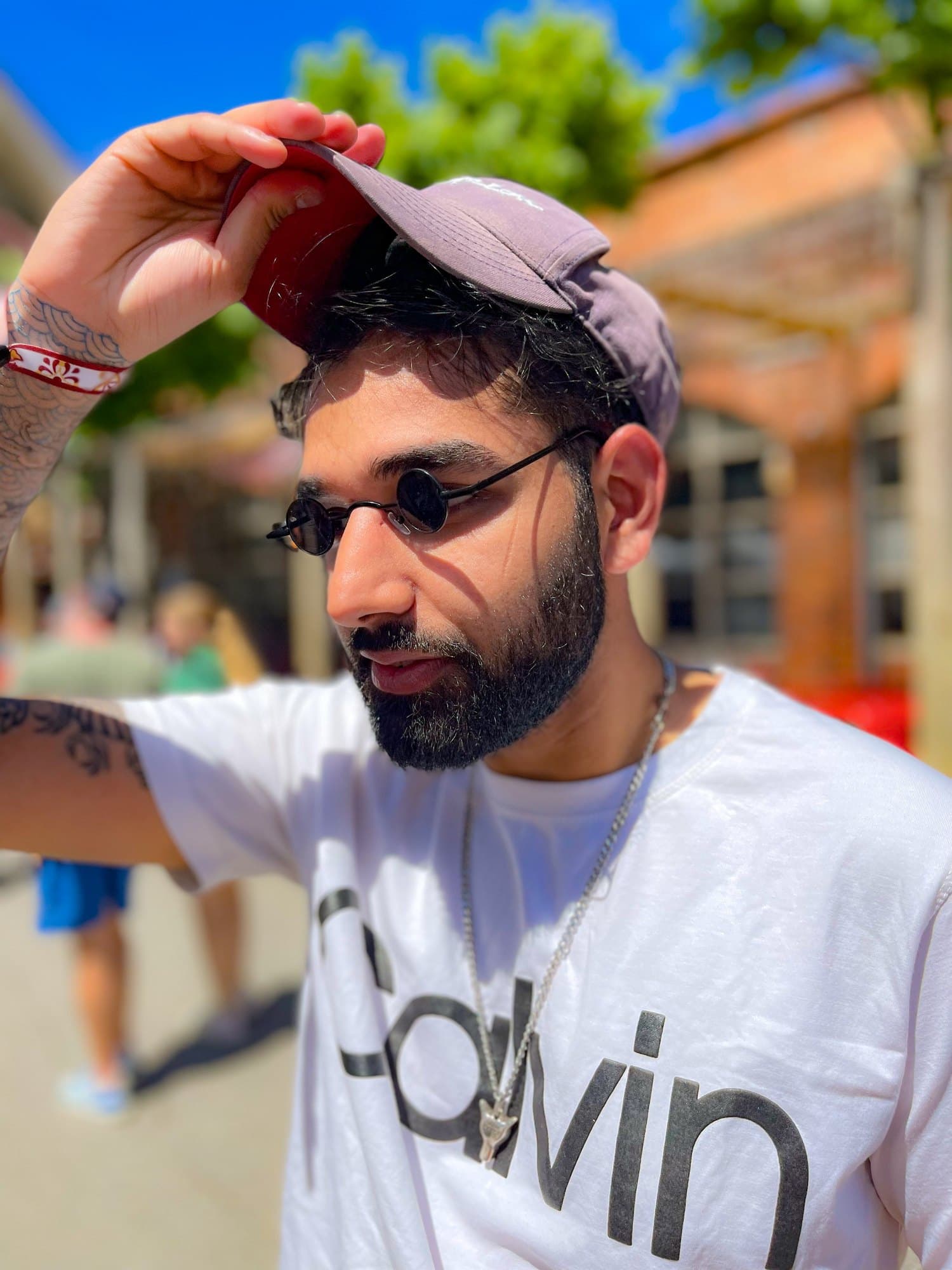 Close portrait of Param in round sunglasses, one hand on the peak of his cap, in bright sun