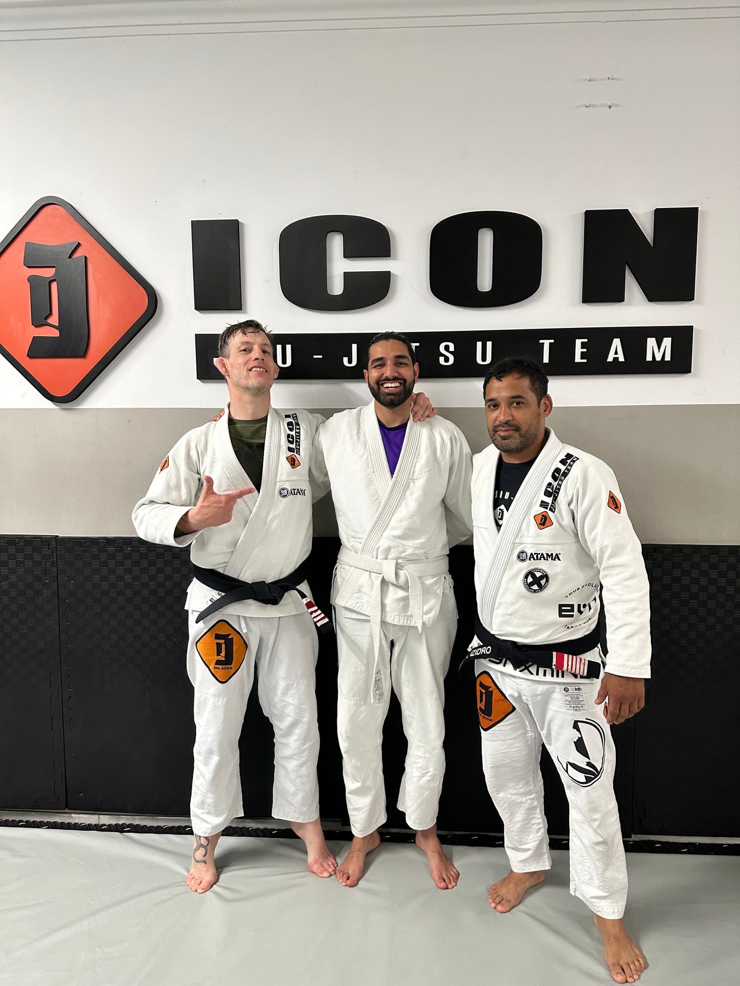 Param in a white gi standing on the mats between two black belts in front of a gym wall sign