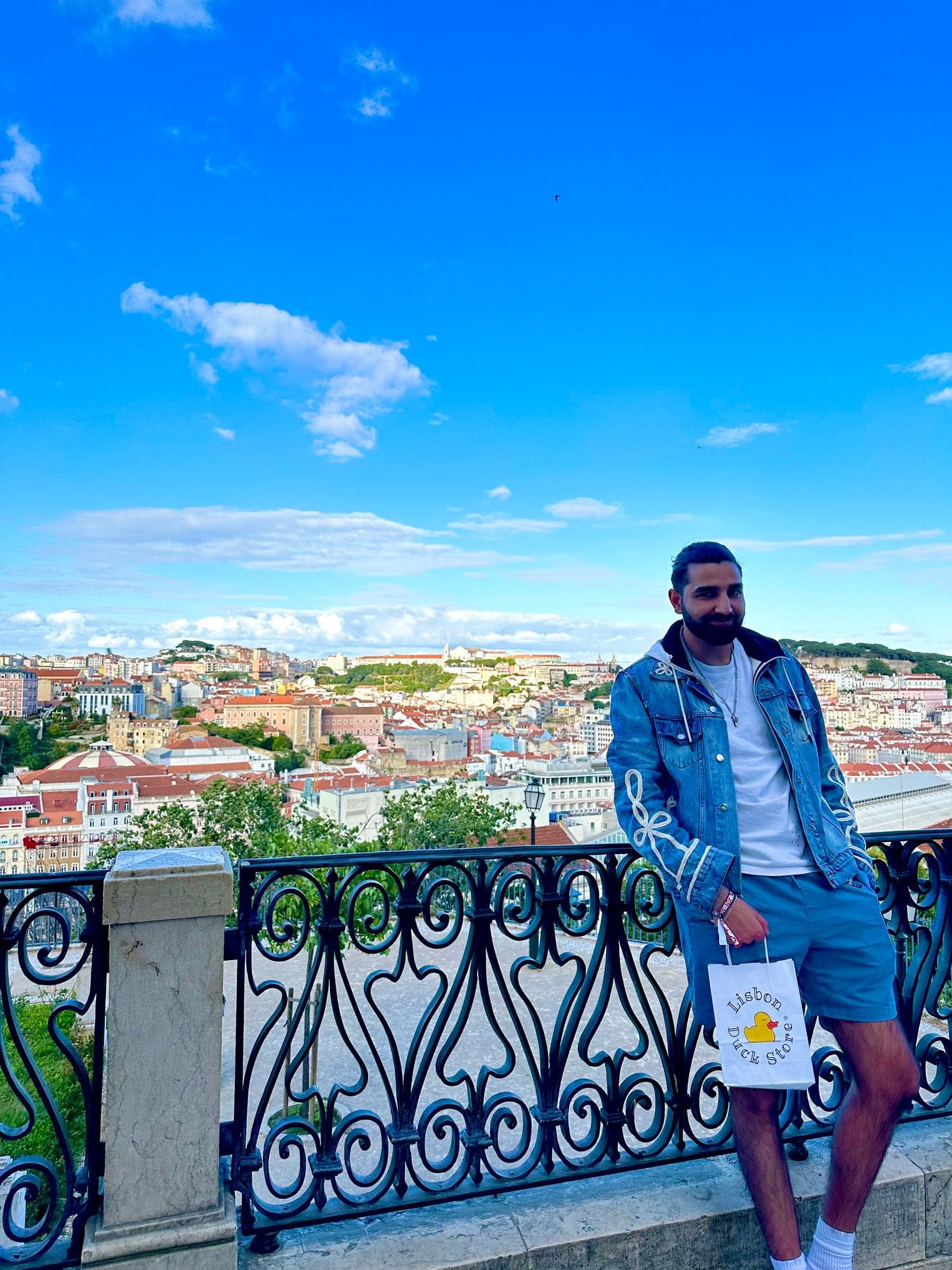 Param in a denim jacket leaning on an ornate iron railing, the city and the castle hill behind him