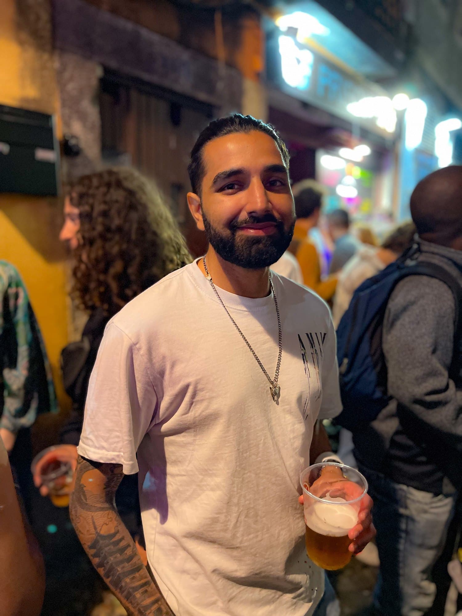Param smiling in a white T-shirt with a pint in his hand on a crowded street at night