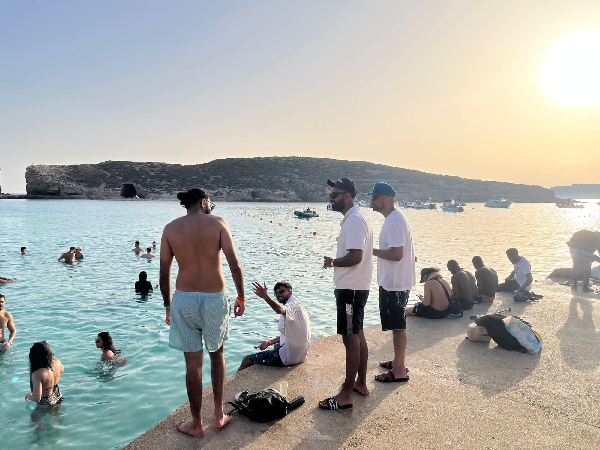 Me from behind, shirtless in pale shorts, at the edge of a stone jetty above turquoise water at sunset, others sitting and standing along the jetty