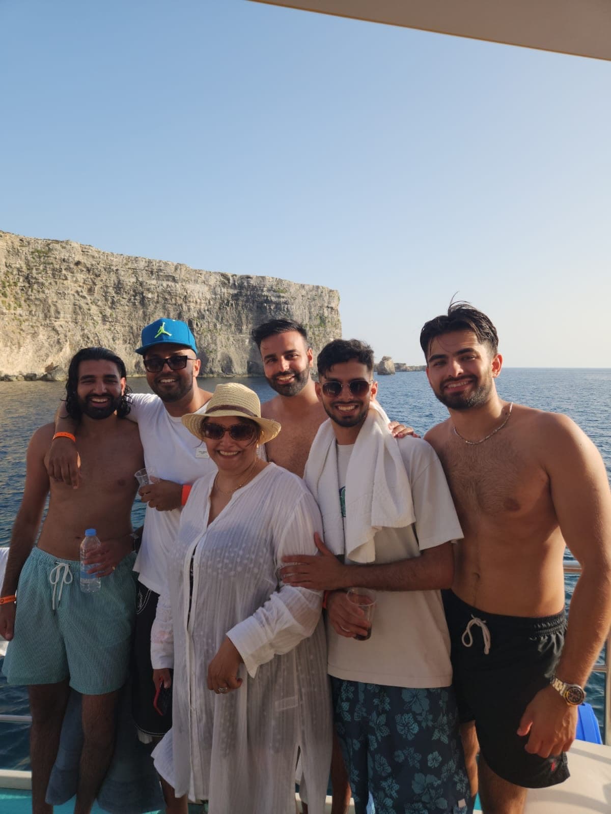 Six of us on a boat deck in front of a pale limestone cliff, me on the far left in striped swim shorts, an older woman in a sun hat in the middle