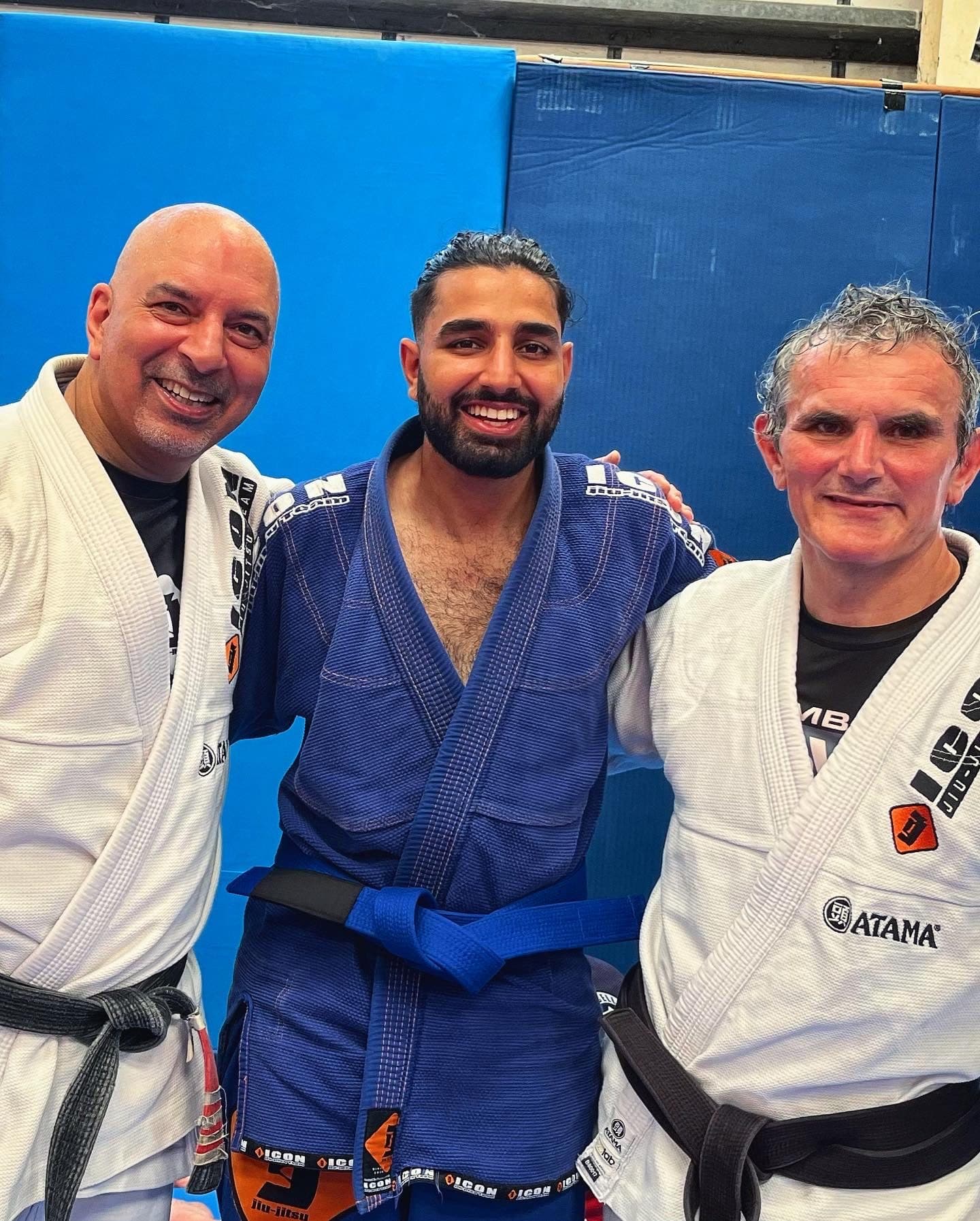 Param, in a blue gi and a new blue belt, smiles between two black-belt coaches in white gis against a padded blue wall