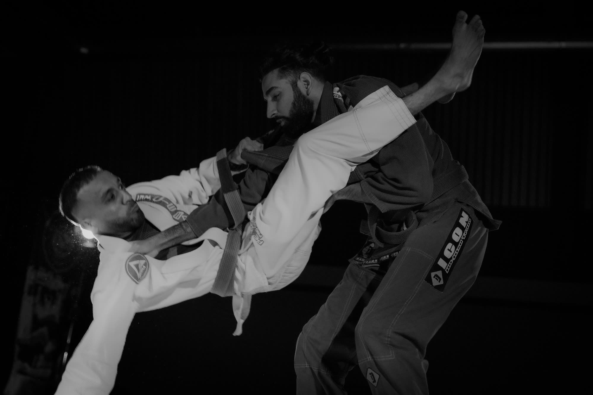 Black-and-white action photograph of Param in a dark gi standing as his opponent in a white gi holds his collar and jumps guard, one leg already up around him