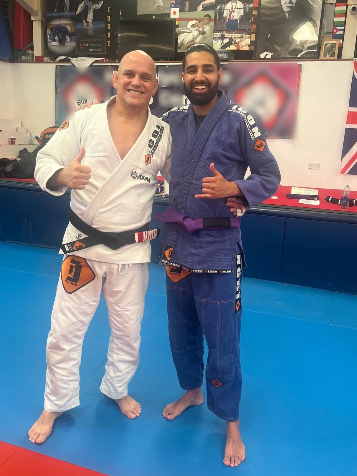 Param, in a blue gi and a new purple belt, stands beside his coach in a white gi and black belt, both grinning, the coach giving a thumbs up