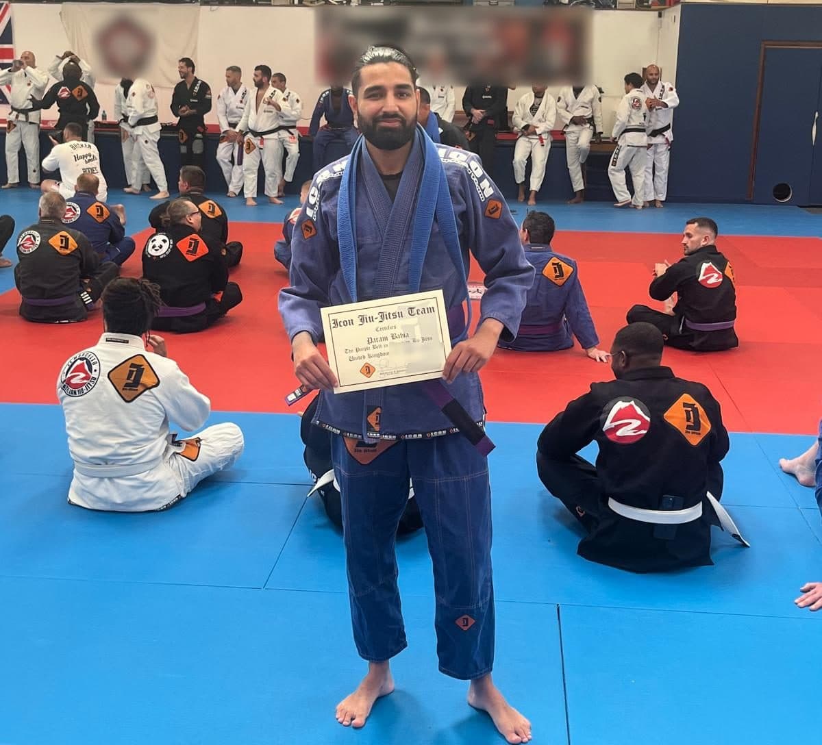 Param, in a blue gi with a purple belt, stands on the mat holding his grading certificate while the rest of the class sits behind him