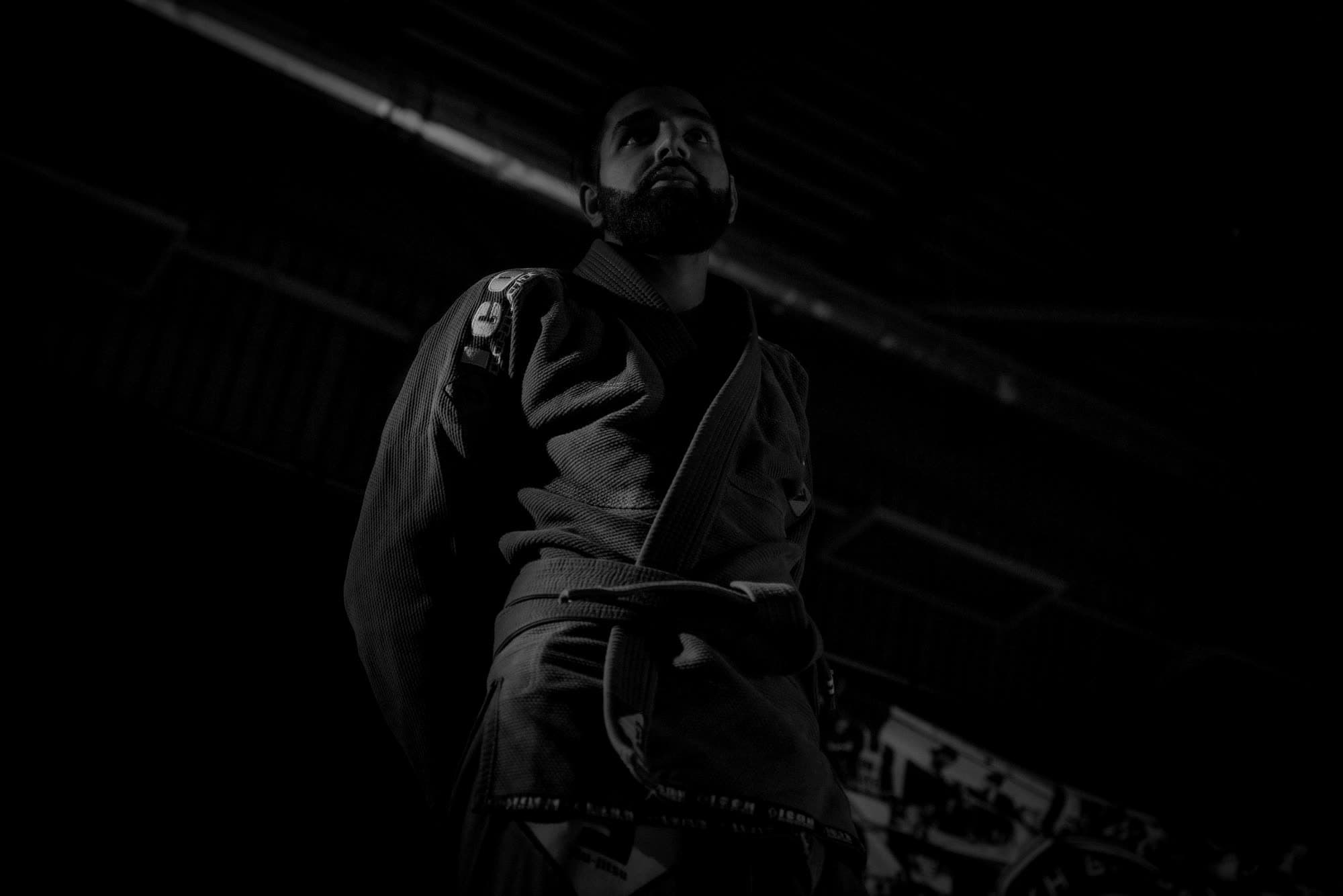 Black-and-white low-angle photograph of Param in a dark gi with his belt tied, looking up and to the left under the hall lights
