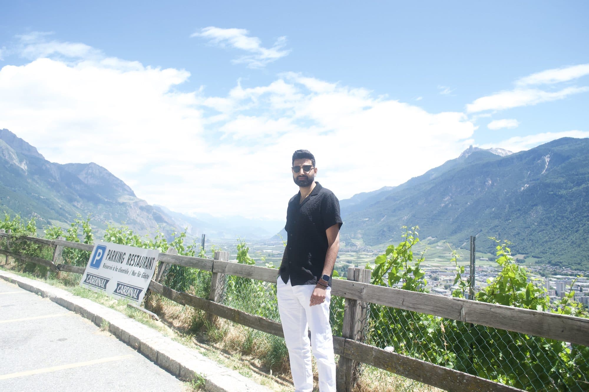 Me in a black shirt and white trousers at a wooden fence above a wide valley, vines and a town below