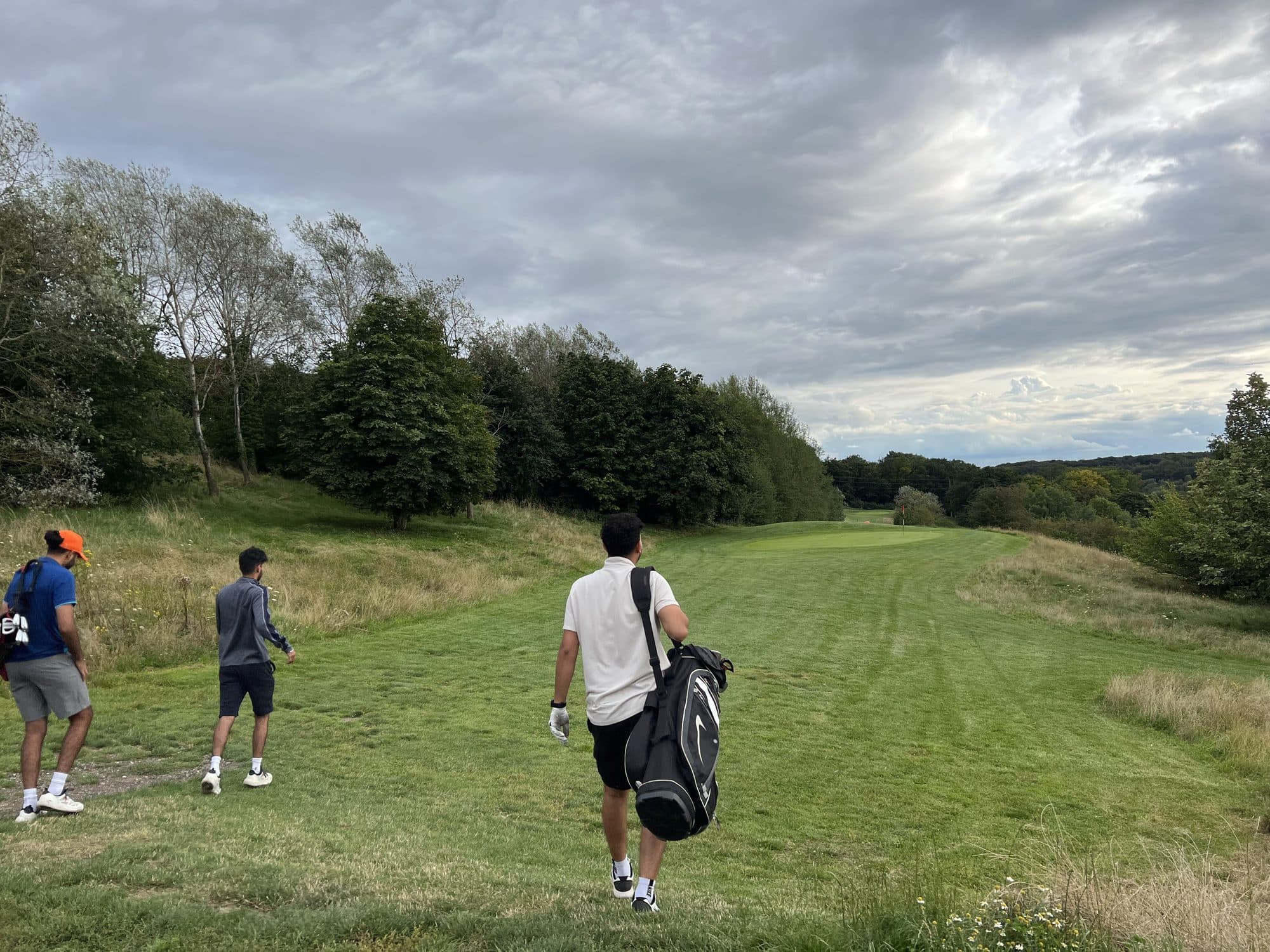 Three golfers seen from behind walking down a fairway towards a distant green, one carrying a bag over his shoulder, trees on the left and grey cloud overhead