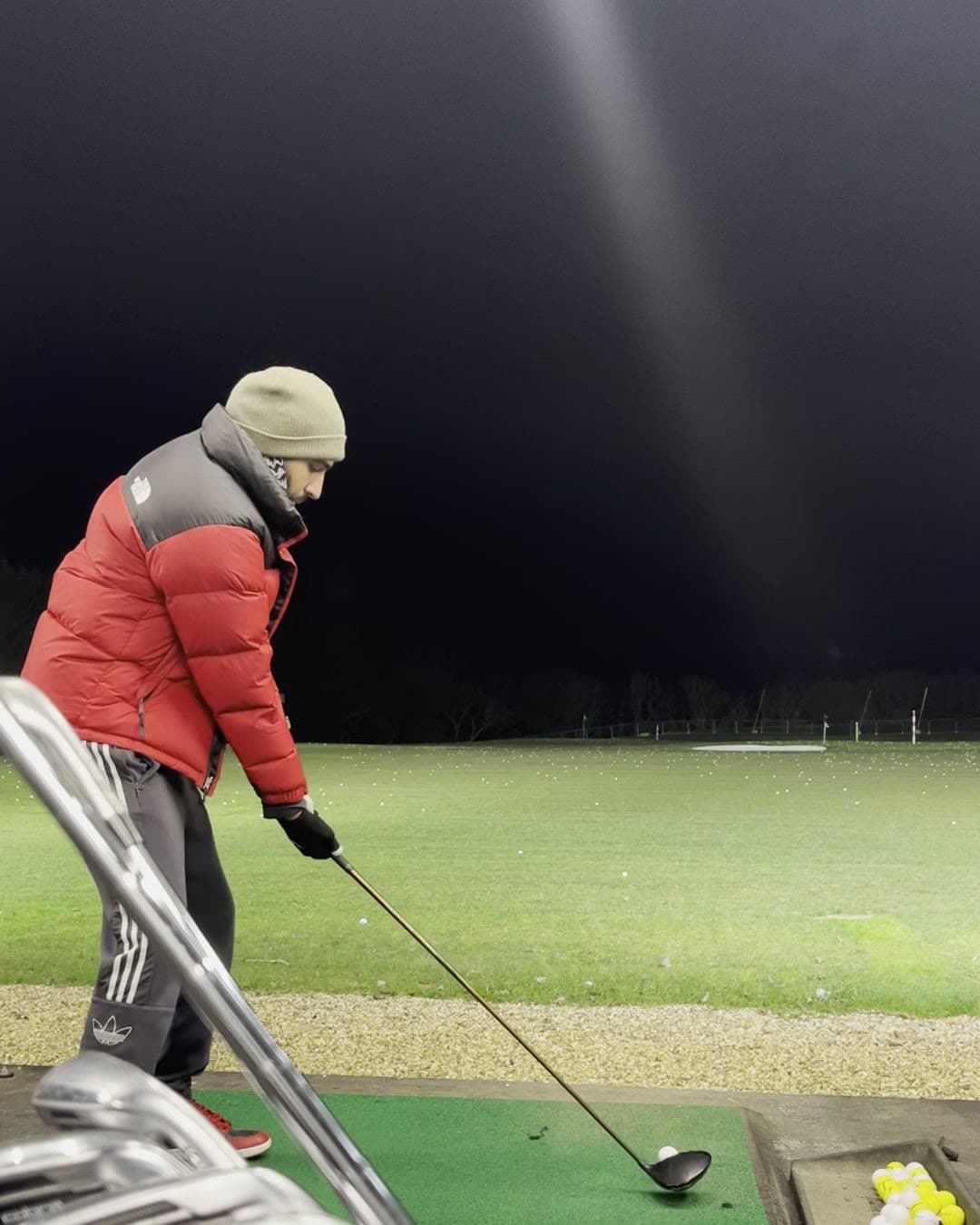 Param in a red puffer jacket and beanie at address with a driver on a floodlit range mat, a bag of irons in the foreground