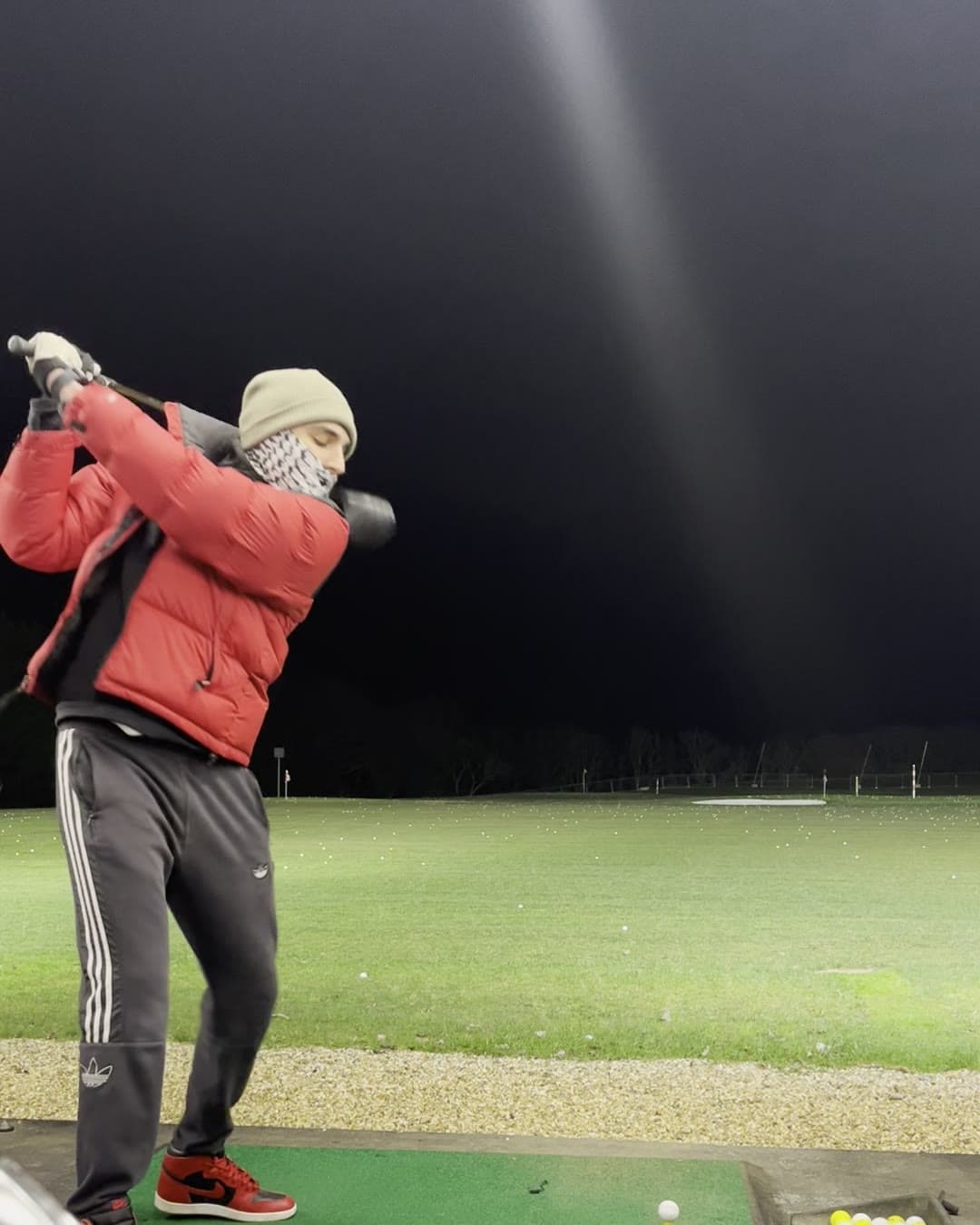 Param at the top of his backswing under the floodlights, scarf pulled up over his chin, the ball still on the tee peg in front of him